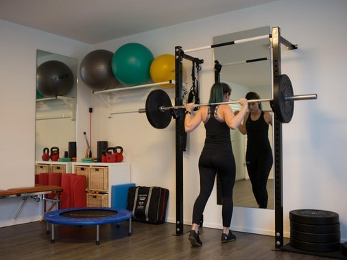 Frau trainiert mit einer Langhantel im Rack, im Hintergrund Gymnastikbälle, Kettlebells und ein Minitrampolin.