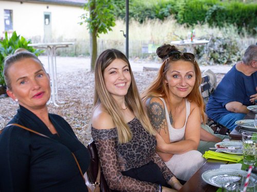 Drei lächelnde Kolleginnen sitzen nebeneinander im Freien am Tisch.