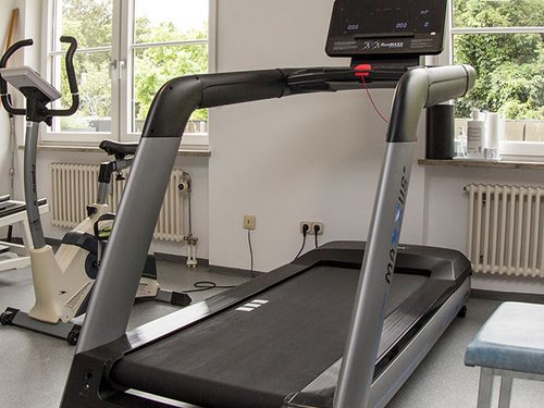 Trainingsraum mit Laufband, Fahrradergometer, Wanduhr und blauem Hocker.