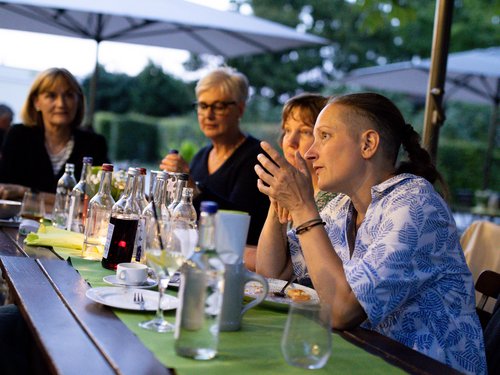 Kolleginnen sitzen gesellig zusammen im Biergarten, im Hintergrund weitere Gäste und ein Gebäude mit türkisen Fensterläden.