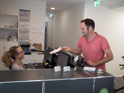 Mitarbeiterin am Empfangstresen reicht einem Patienten ein Dokument, im Hintergrund ein Rollator.