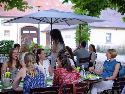 Kolleg:innen sitzen gesellig an einer langen Tafel unter einem Sonnenschirm im Garten des Hofguts.