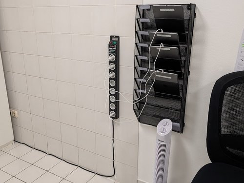 Steckdosenleiste und Zeitschriftenhalter mit Tablets an einer gefliesten Wand, daneben ein Ventilator und Bürostuhl.
