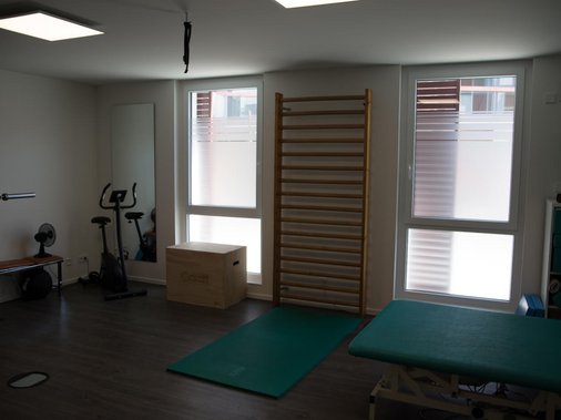 Kleiner Behandlungsraum mit Liege, Hocker und Fenster.