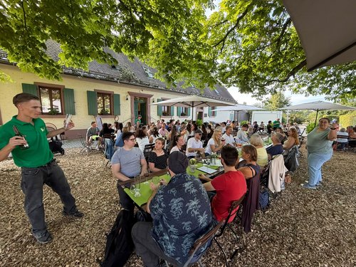 Kolleg:innen sitzen und stehen bei geselligem Beisammensein vor dem Gebäude des Hofguts im Schatten großer Bäume.