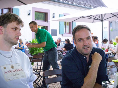 Kolleg:innen unterhalten sich an einem Tisch im Biergarten, im Hintergrund bedient ein Kellner weitere Gäste.