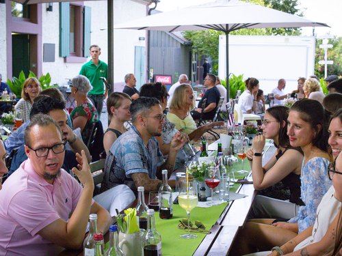 Gruppe sitzt gesellig zusammen an einer langen Tafel unter einem Sonnenschirm.