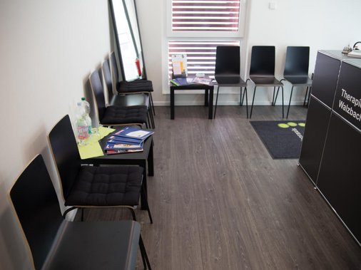 Wartebereich mit Sitzreihe, Beistelltisch mit Broschüren und Empfangstresen im Hintergrund.