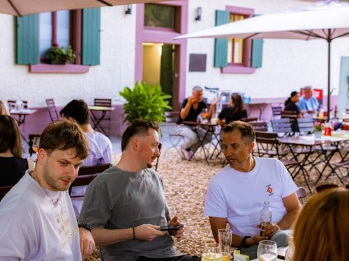 Gruppe von Kolleg:innen sitzt bei einem Getränk zusammen im Biergarten des Hofguts, im Hintergrund weitere Tische und ein pastellfarbenes Gebäude mit türkisen Fensterläden.