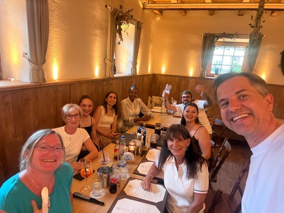 Große Gruppe von Kolleg:innen sitzt lächelnd am Restauranttisch, ein Mitarbeiter macht ein Gruppenselfie.