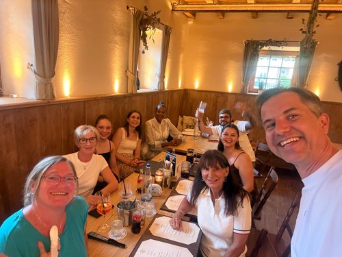 Große Gruppe von Kolleg:innen sitzt lächelnd am Restauranttisch, ein Mitarbeiter macht ein Gruppenselfie.