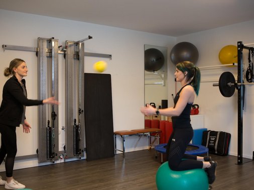 Zwei Frauen üben Ballwurf-Koordination, eine kniet auf einem Gymnastikball.