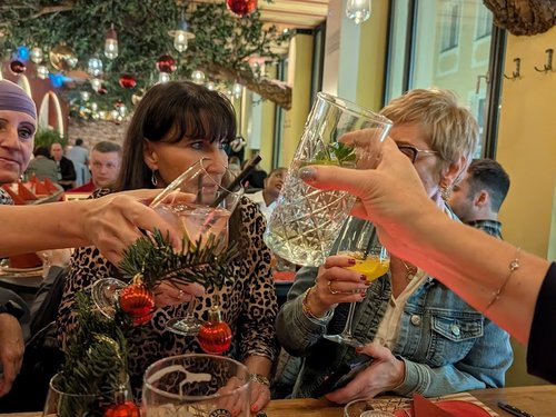 Kolleginnen stoßen mit weihnachtlich dekorierten Cocktailgläsern an, im Hintergrund Weihnachtskugeln von der Decke.