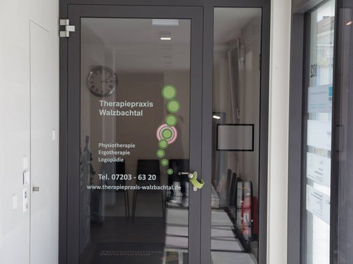 Eingangstür der Praxis mit Beschriftung „Therapiepraxis Walzbachtal" und Wanduhr im Innenbereich.